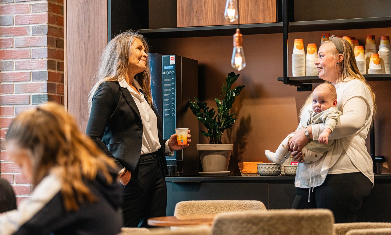 Två kvinnor står vid kaffeautomat varav den ena håller i en bebis