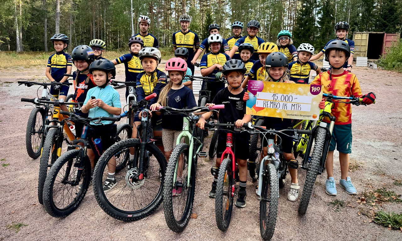 Ett stort grupp med barn och ledare står med sina cyklar på en grusplan. En av barnen håller upp vinstchecken på 15 000 kr.