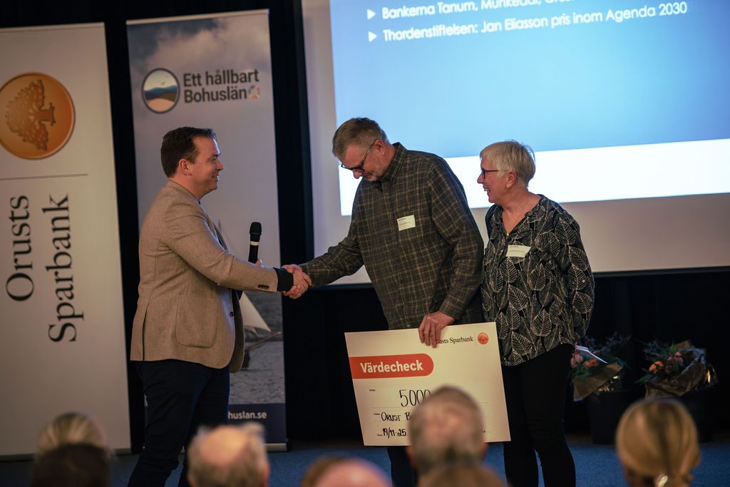 En representant från Orusts Sparbank överlämnar en värdecheck till två mottagare på en scen under ett evenemang.