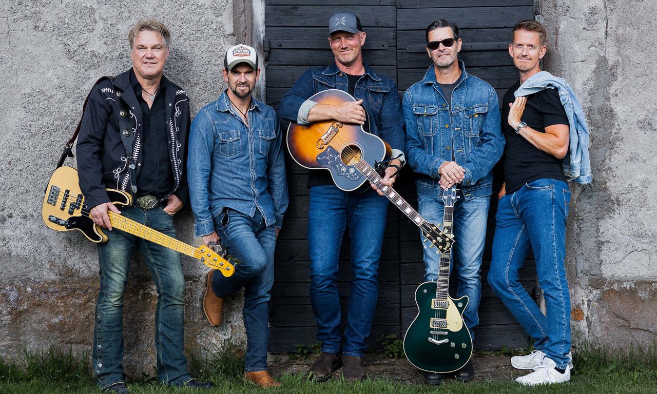 Fem män står mot en ladugårdsvägg och tre håller i en gitarr