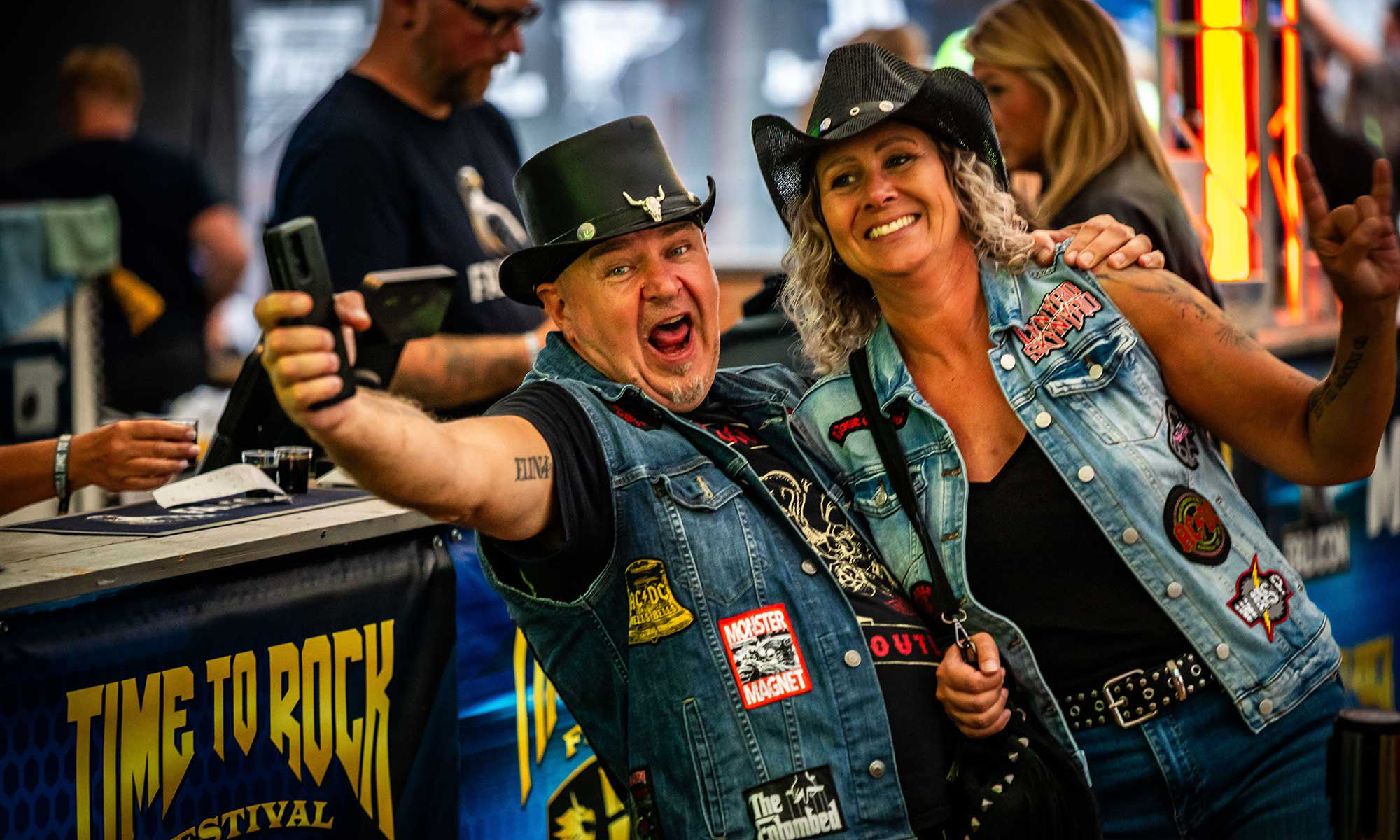 Två vuxna i jeansvästar poserar för en selfie på en livlig rockfestival. De står framför en bardisk. Synlig text inkluderar "TIME TO ROCK FESTIVAL" på en banderoll bakom dem..
