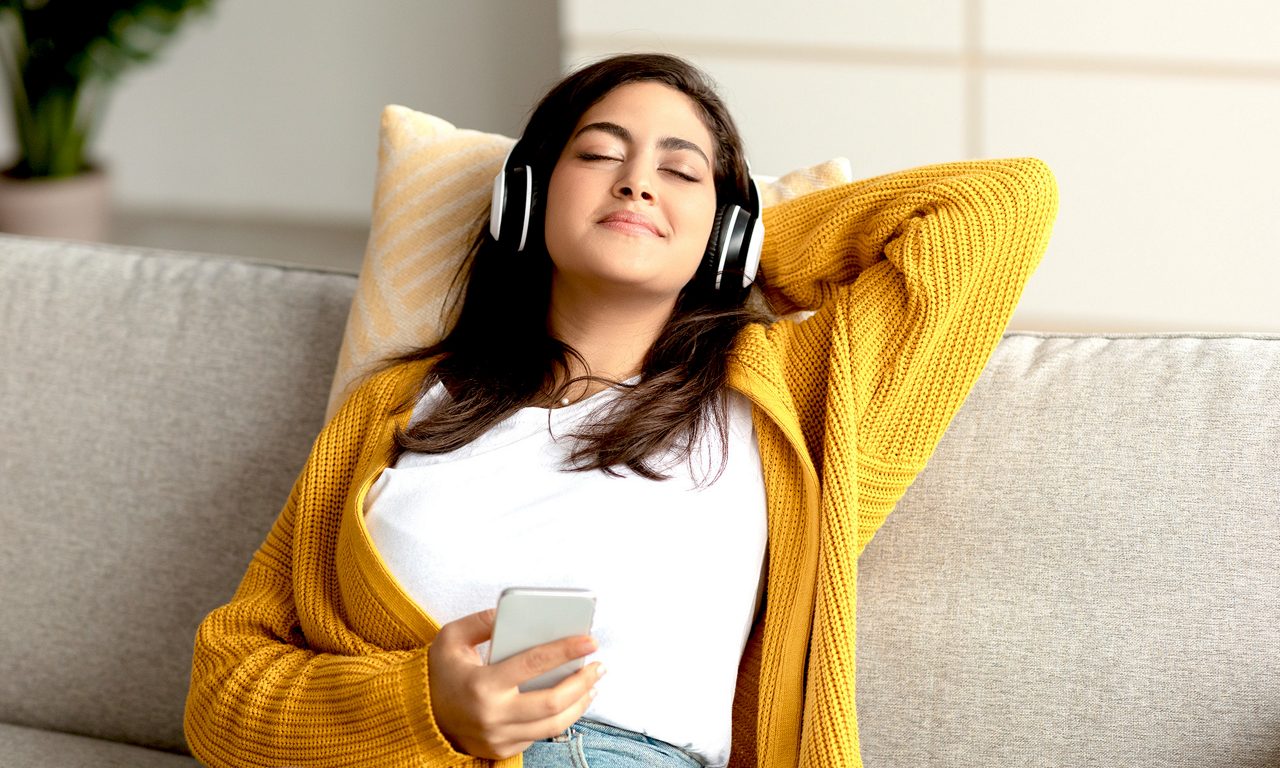 En ung tjej med hörlurar sitter tillbakalutad i en soffa med slutna ögon