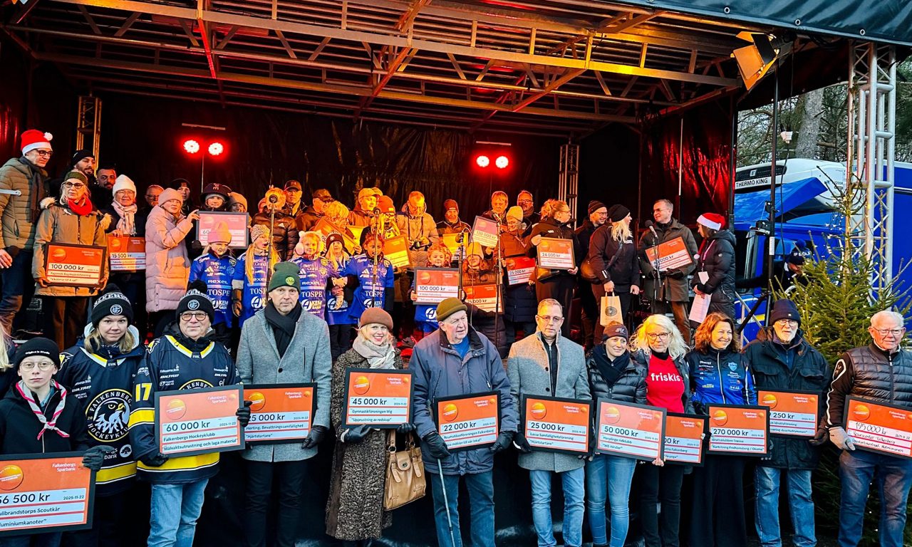 Stort antal bidragstagare poserar med checkar på scen vid utomhusutdelning i vintermiljö.