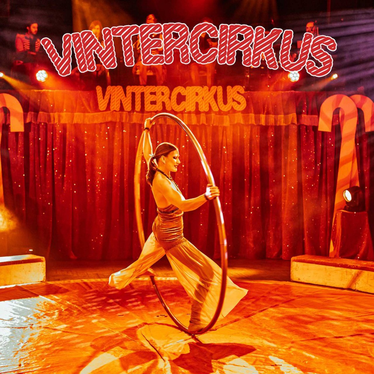 A female circus performer spins inside a large metal hoop on a brightly lit stage. The scene takes place in a winter-themed circus tent with red curtains and festive decorations.