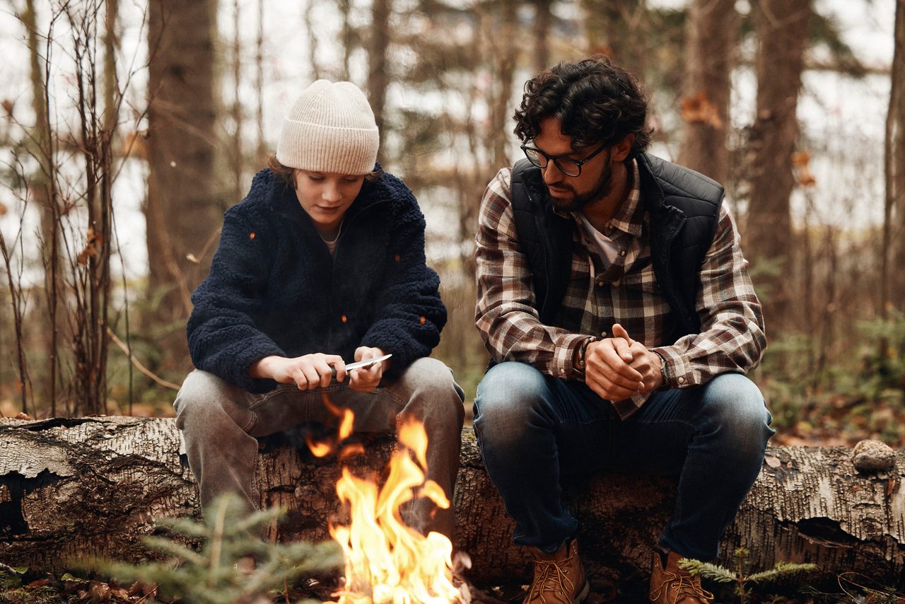 En vuxen och ett barn sitter tillsammans i skogen vid en liten eld, omgivna av träd och natur.