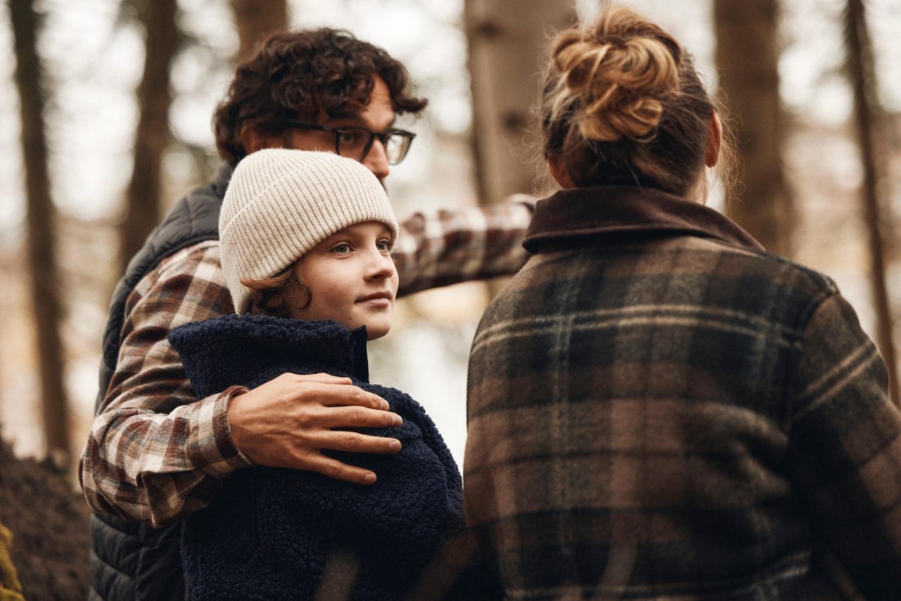 Två vuxna och ett barn går tillsammans i skogen, där en vuxen skyddande håller armen om barnet.