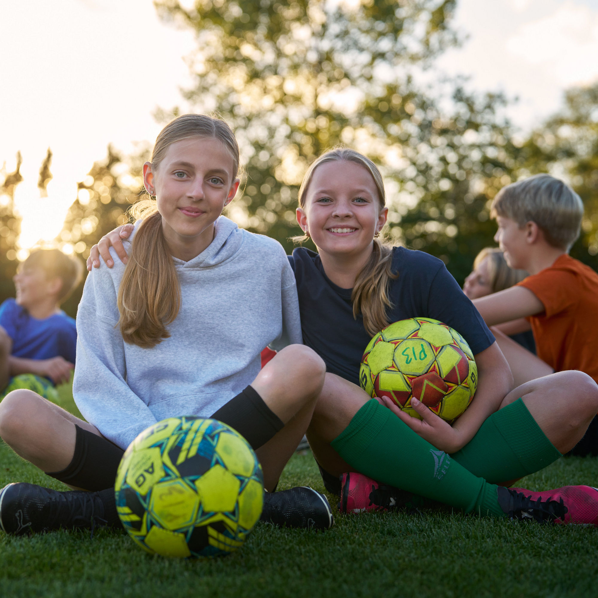 Unga fotbollsspelare sitter i gräset och två tjejer håller om varandra. 