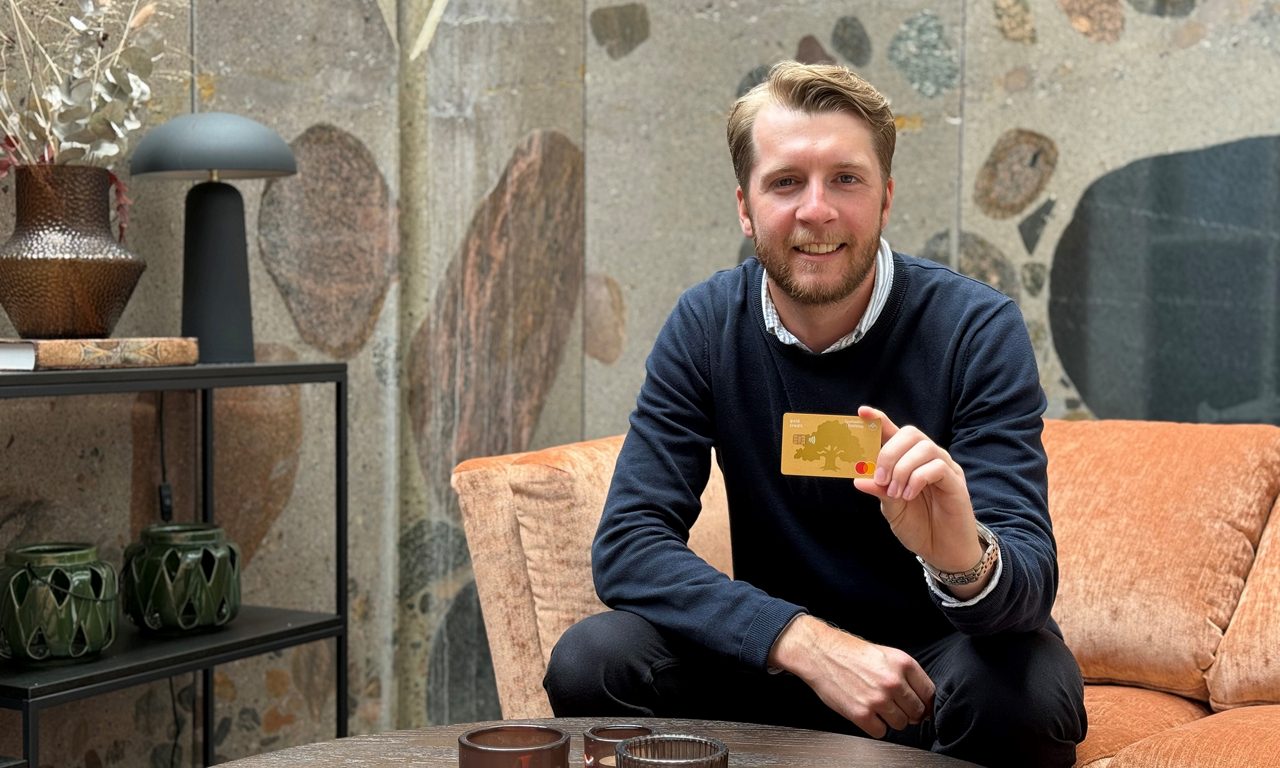 man i vit skjorta och mörkblå tröja håller ett betalkort i handen och tittar in i kameran