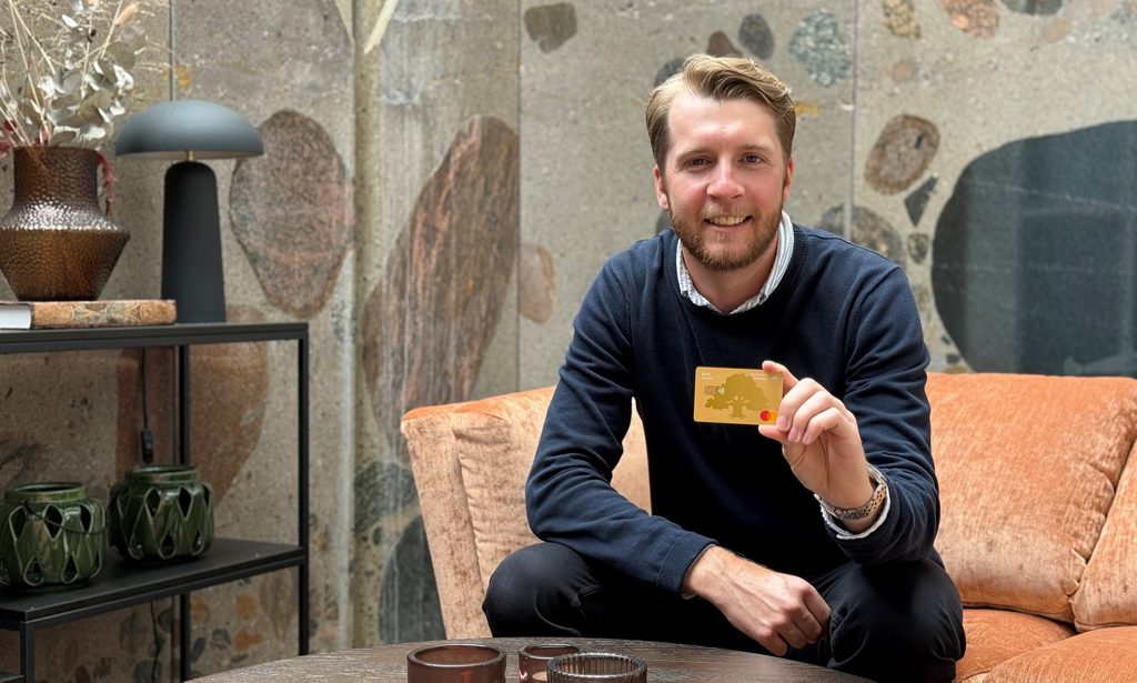 man i vit skjorta och mörkblå tröja håller ett betalkort i handen och tittar in i kameran