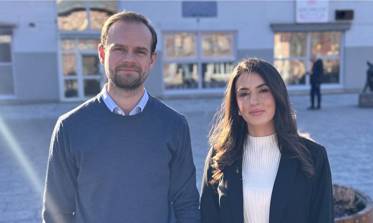 en man och kvinna står bredvid varandra med ryggen till bankkontorets entré och tittar in i kameran