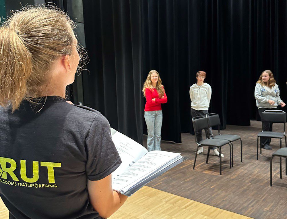 En kvinna som håller en pärm i ett stort rum med några stolar framför tre ungdomar