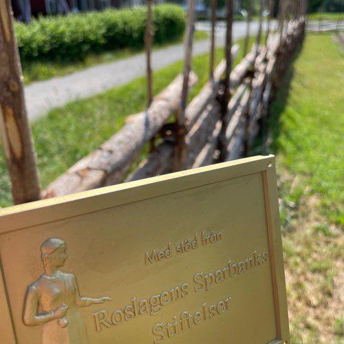 Guldig skylt med texten "Med stöd från Roslagens Sparbanks Stiftelser" framför staket