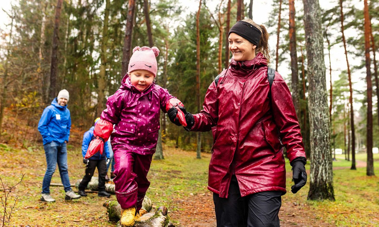 Barn i skogen balanserar på en stock och håller en vuxen kvinna i handen.