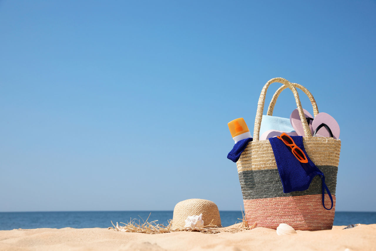 Fylld strandbag med glasögon, flipflop och handduk på en strand med havet i bakgrunden