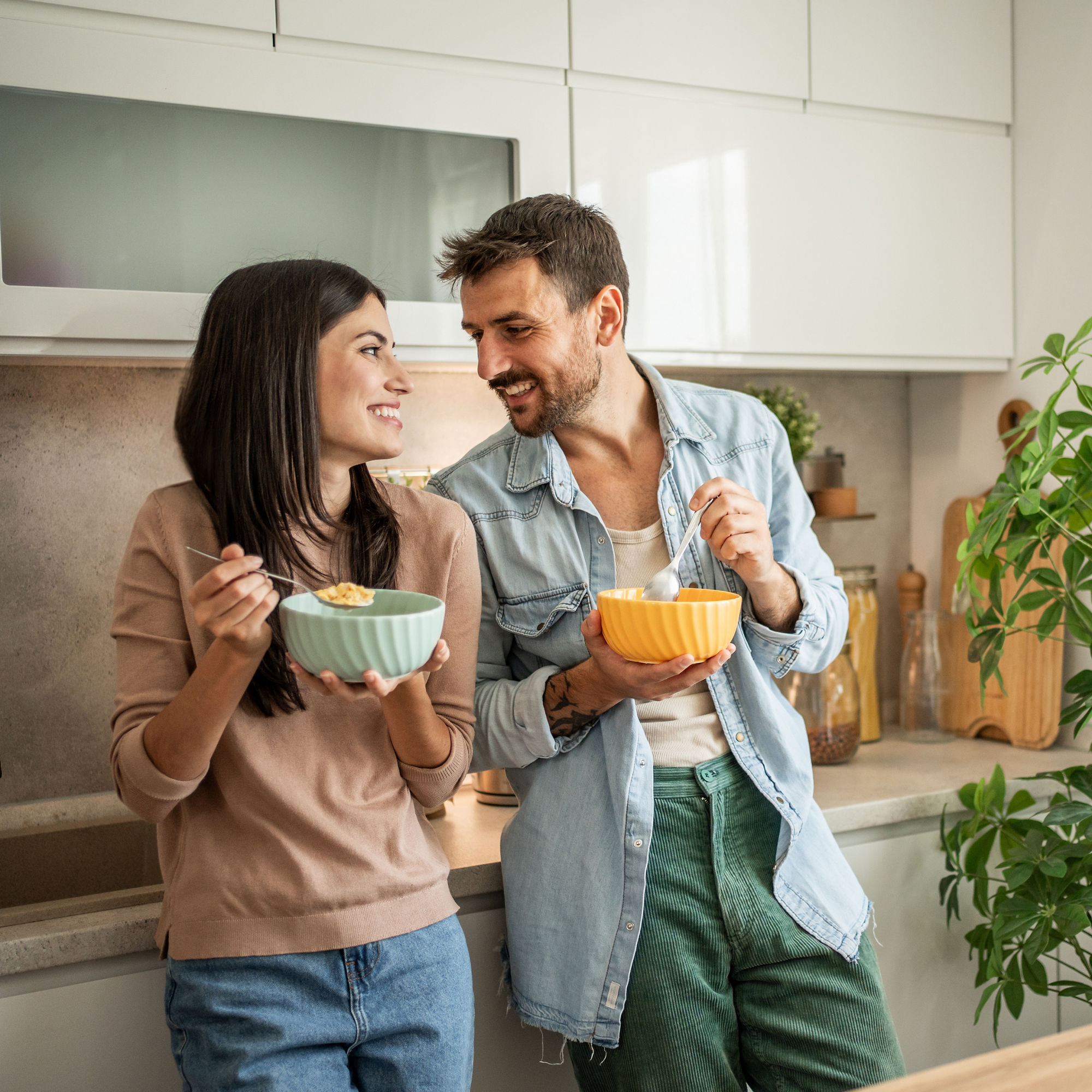 Ett par står i ett ljust kök och äter frukost ur skålar samtidigt som de ler och pratar med varandra, med en grön krukväxt i bakgrunden.