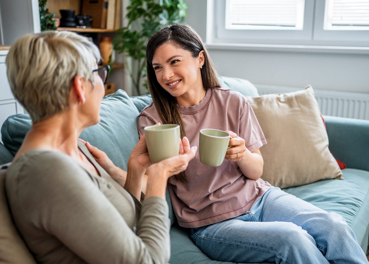 Två kvinnor sitter i en soffa och ler mot varandra medan de håller varsin kopp te eller kaffe, i en varm och avslappnad hemmiljö.