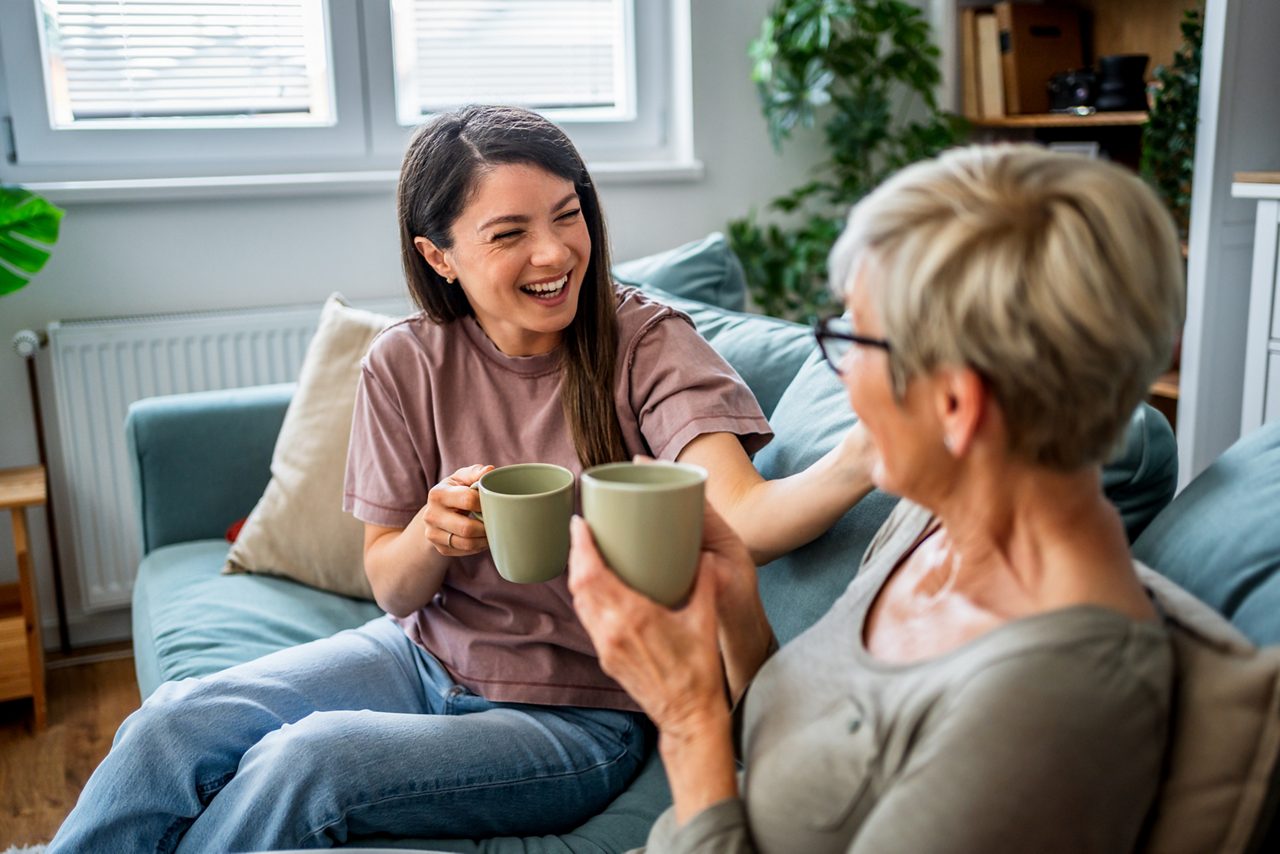 En glad mamma och dotter njuter av en avkopplande stund tillsammans, skrattar och samtalar över en kopp kaffe i soffan i sitt mysiga vardagsrum.