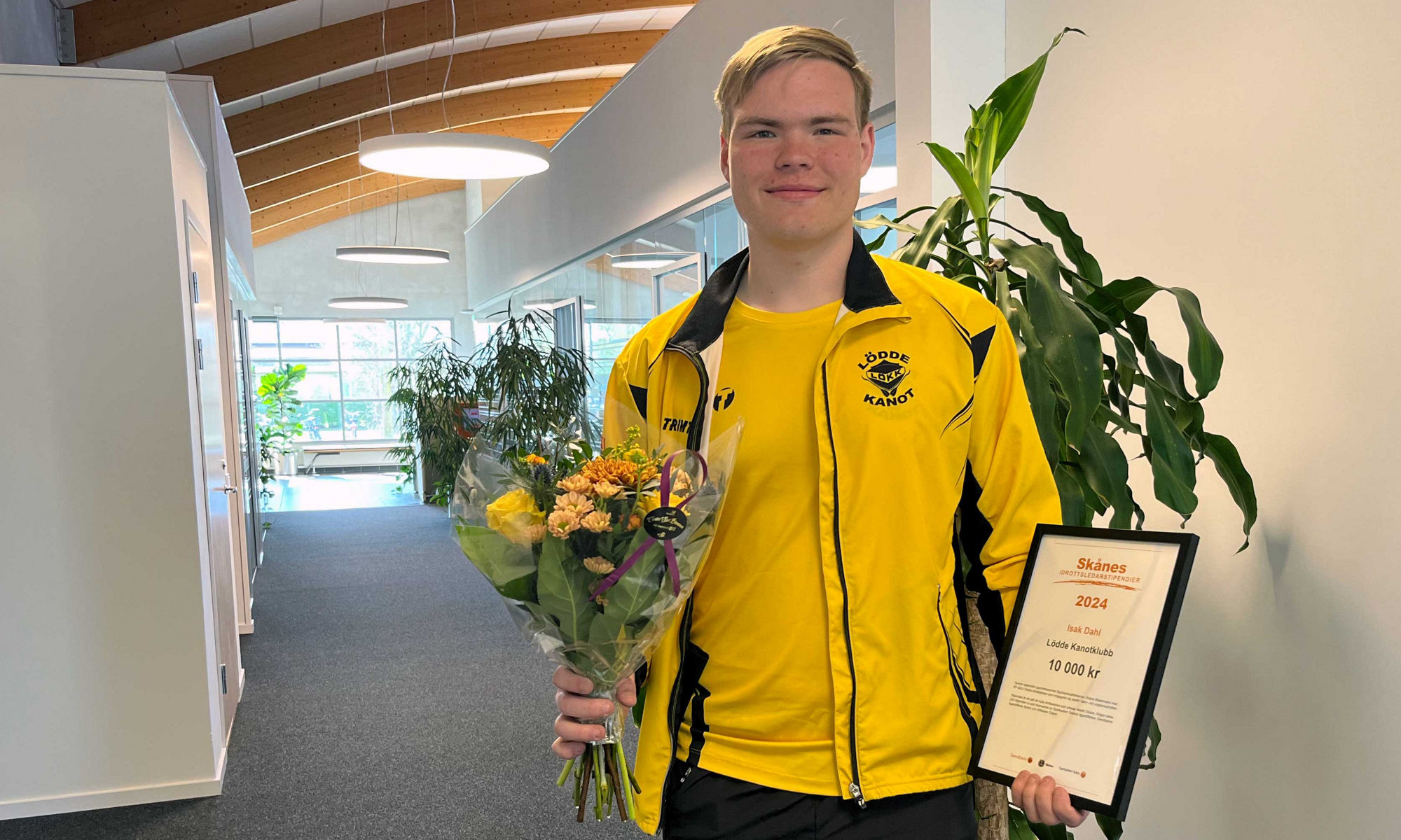 Isak Dahl håller sitt Skånes Idrottsledarstipendium och blommor på bankkontoret.