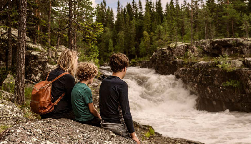 Tre personer sitter på stenig mark med utsikt över en kraftfull flod eller ett vattenfall i en skogsmiljö. Gruppen, troligen en förälder och två barn, vänder sig bort från kameran medan de tittar på det forsande vita vattnet. Höga vintergröna träd och karga klippor omger scenen och skapar en naturlig, avlägsen atmosfär. En brun ryggsäck sticker ut mot landskapets jordnära toner. Den övergripande stämningen känns fridfull, äventyrlig och kopplad till naturen.