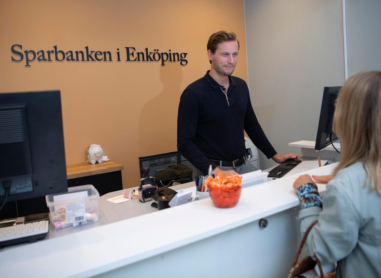a man in a black shirt is standing next to a computer looking at a customer on the other side of the counter. Sparbanken i Enköping is written on the wall behind him.