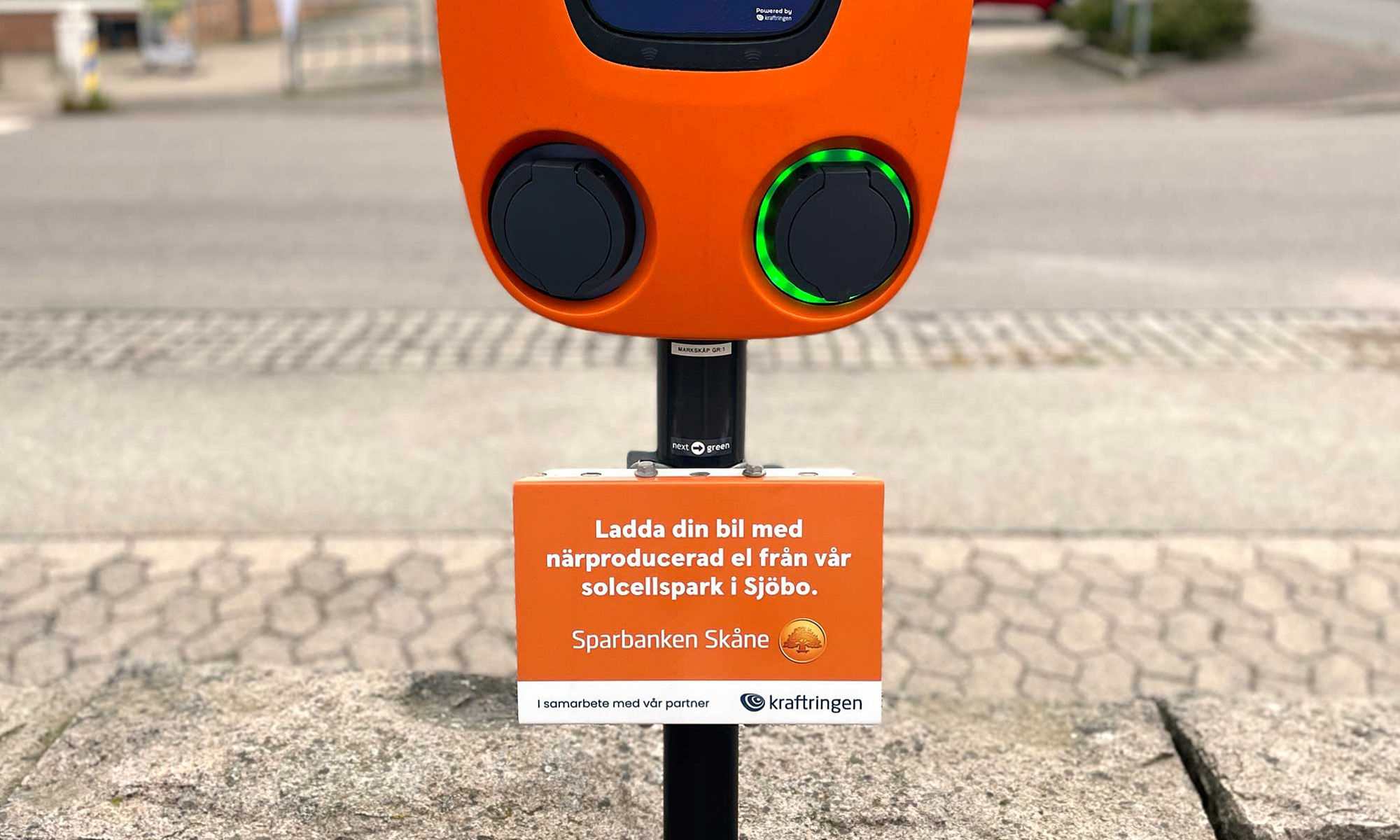 En orange laddstolpe med en skylt på med texten "Ladda din bil med närproducerad el från vår solcellspark i Sjöbo".