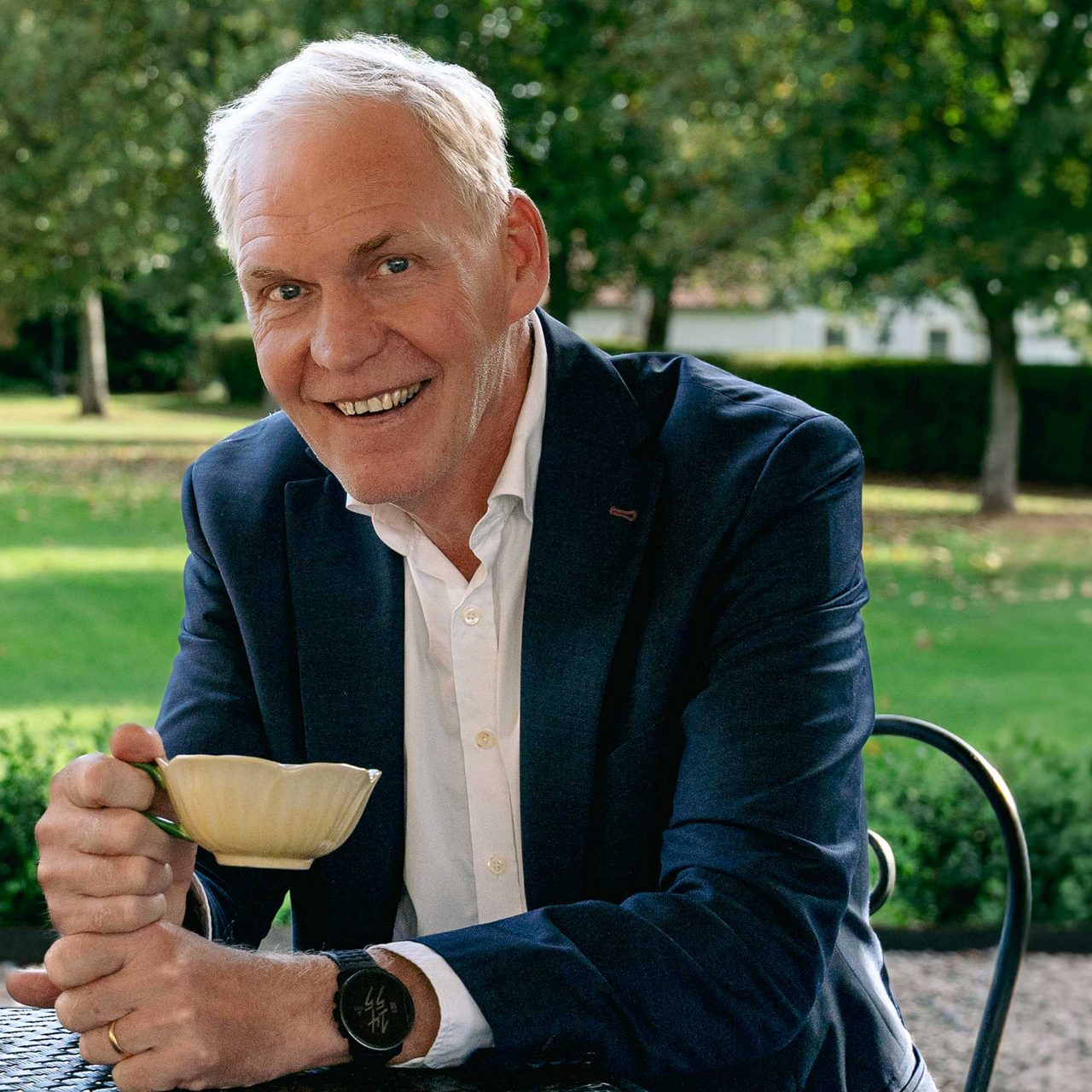 en man i blå kavaj och vit skjorta sitter i en trädgård och håller en kaffekopp i handen