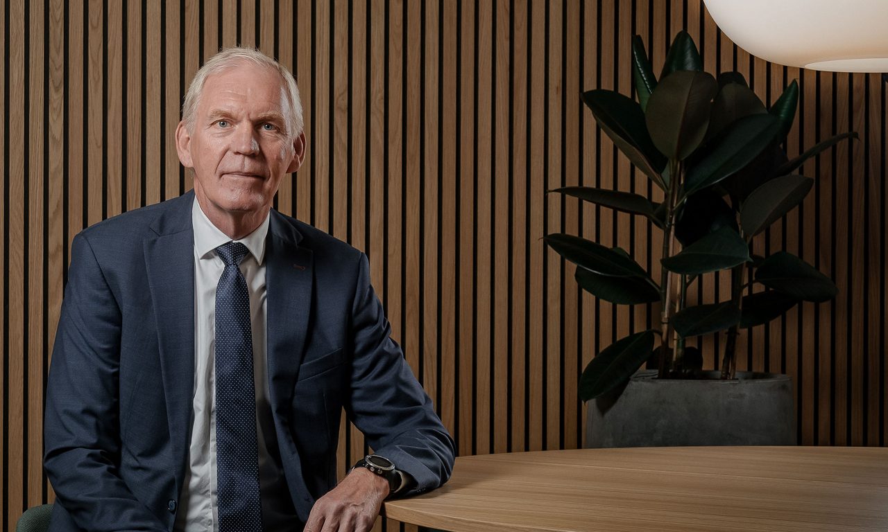 man i kostym och slips står vid ett bord och ser in i kameran