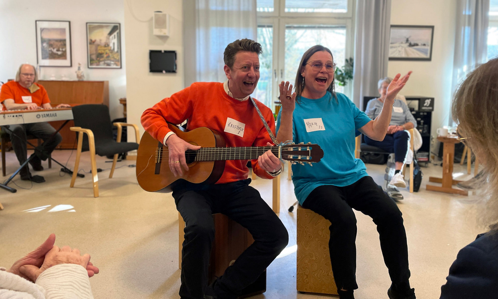 En man och en kvinna sitter och sjunger och spelar gitarr  framför deltagare på musikträff.