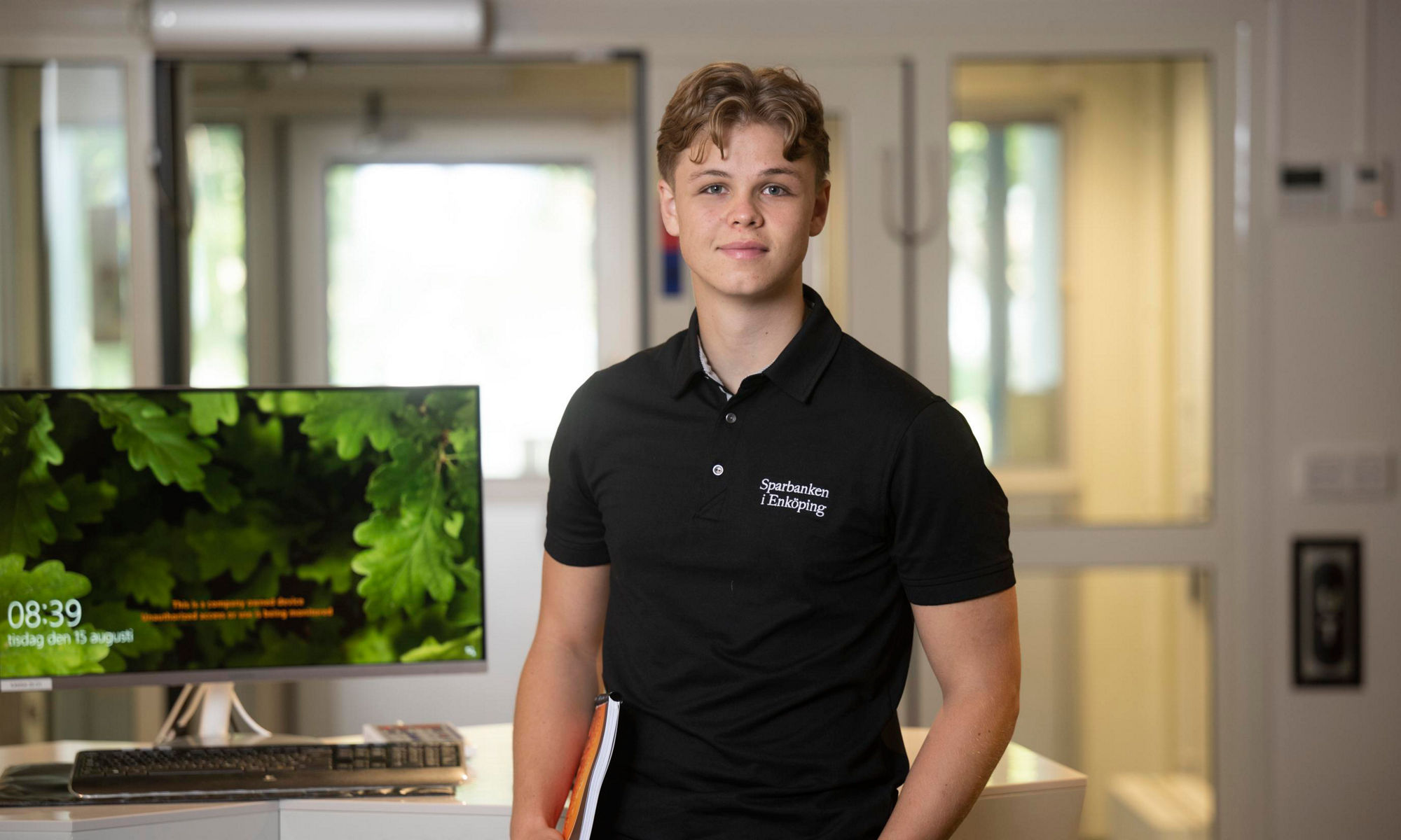 En ung maniklädd svart kortärmad tröja står framför en datorskärm och tittar in i kameran