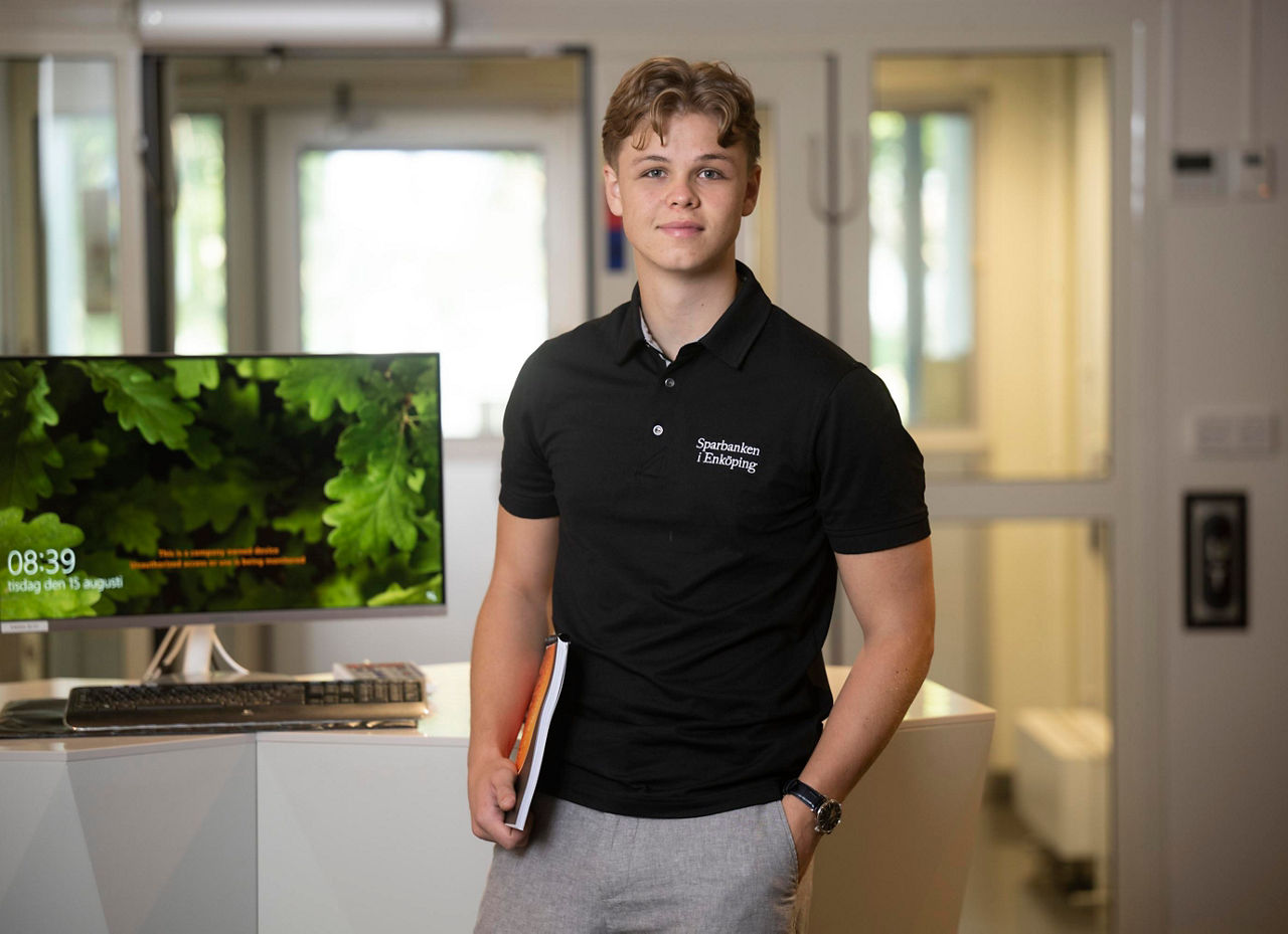 En ung maniklädd svart kortärmad tröja står framför en datorskärm och tittar in i kameran