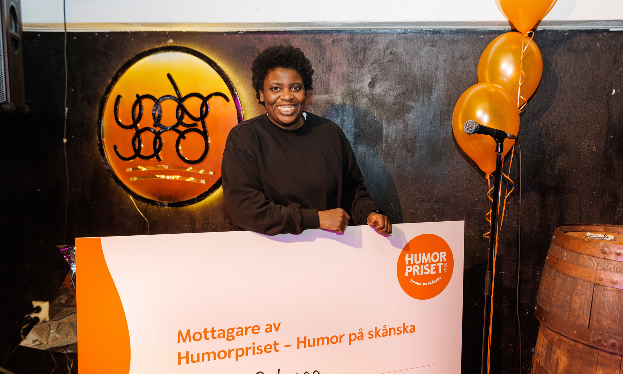 The comedian Petrina Solange stands indoors holding a large check for the Humorpriset award. 