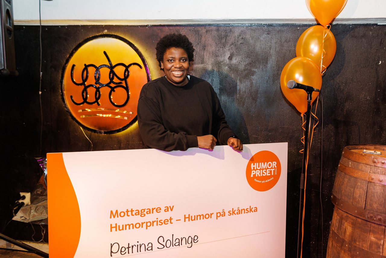 The comedian Petrina Solange stands indoors holding a large check for the Humorpriset award. 