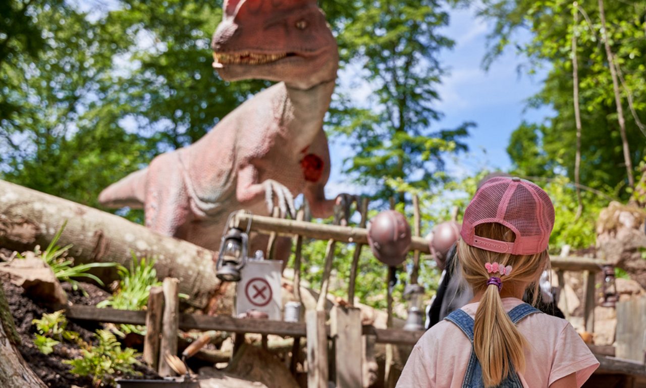 Ett litet barn som bär en rosa keps och hängselbyxor tittar upp på en stor dinosauriefigur i en utomhusutställning.