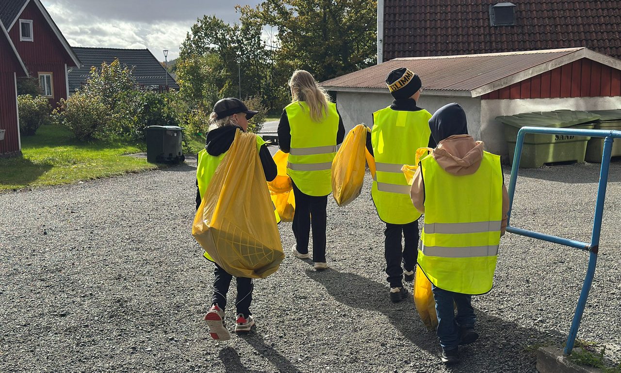En grupp med barn med gula västar på sig går med gula sopsäckar in närheten av röda trähus.
