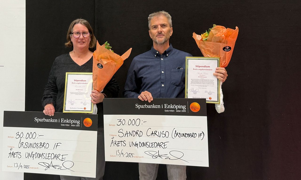 en man i blå skjorta och en kvinna klädd i svart tittar mot kameran, hållandes i varsin stor bankcheck, varsin blombukett samt inramat diplom