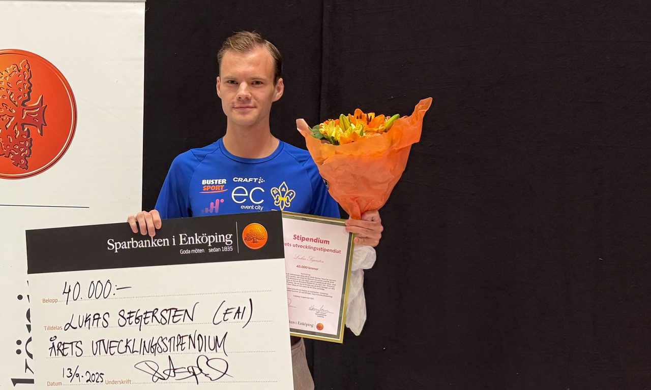 en ung  man i blå tröja tittar mot kameran, hållandes i stor bankcheck, blombukett samt inramat diplom