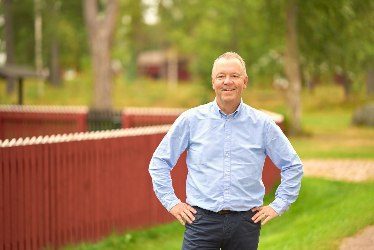 Leende man står framför ett rött staket och gröna träd
