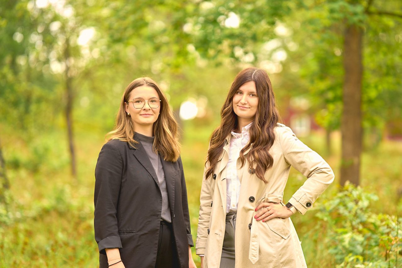 Två kvinnor framför grönskande träd, en solig dag