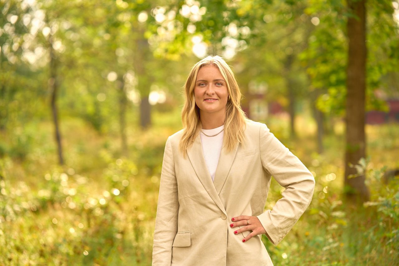 Leende kvinna står framför gröna träd en solig dag