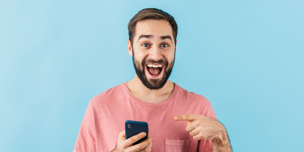 Mann mit kurzen Haaren und Bart hält ein Smartphone in der Hand und lacht begeistert. Er deutet mit dem Zeigefinger der linken Hand auf sein Handy.