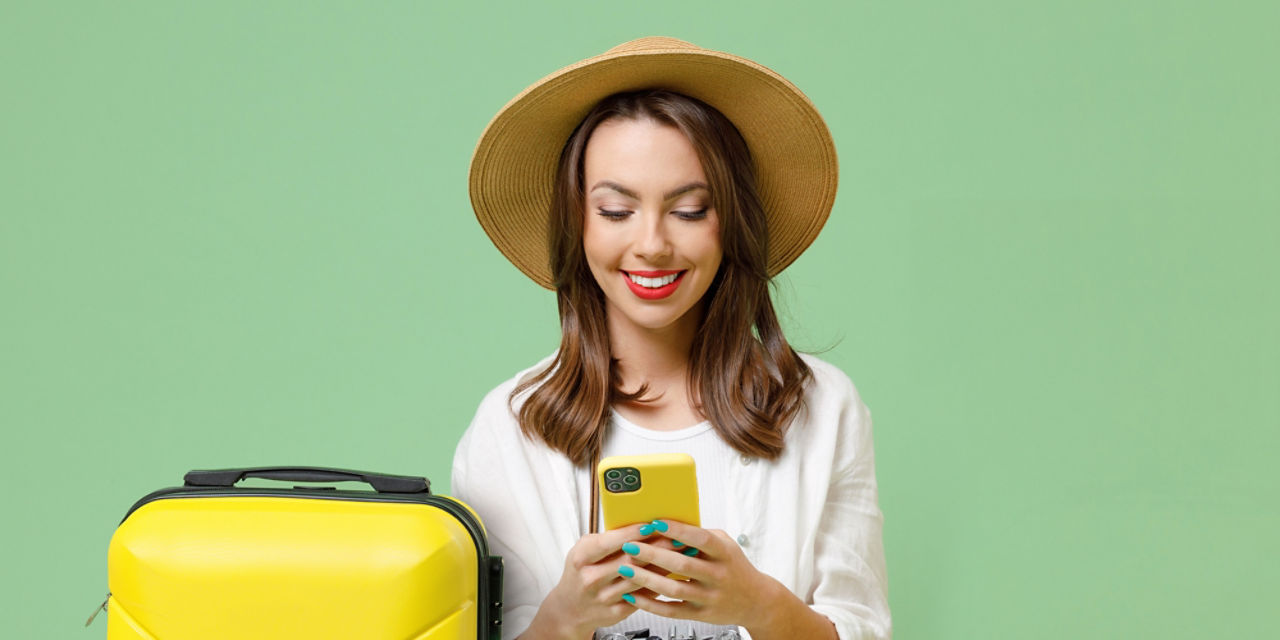 Frau mit Sonnenhut steht neben einem gelben Hartschalenkoffer und sieht lächelnd  in ihr Handy.
