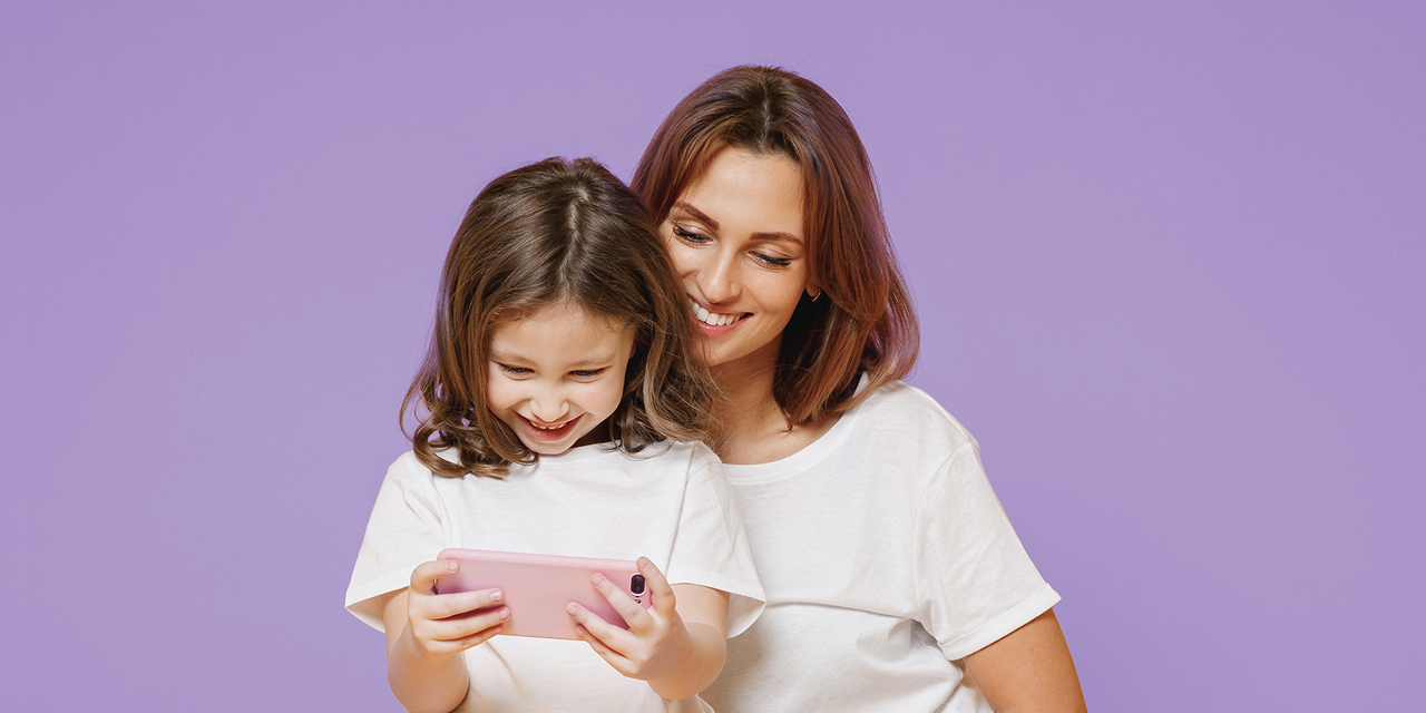Junges Mädchen hält mit beiden Händen ein Smartphone und schaut auf den Bildschirm. Ihre Mutter steht hinter ihr und blickt ihrer Tochter über die Schulter. Beide lächeln.