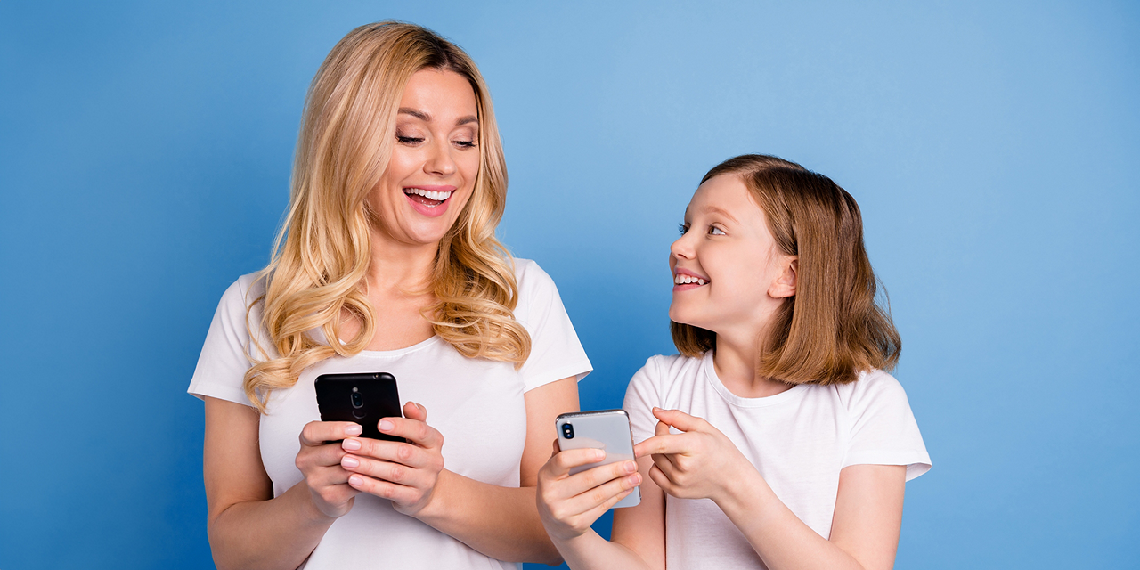Mutter und Tochter halten beide lächelnd ein Smartphone in der Hand. Tochter zeigt Mutter etwas auf Ihrem Handy und Mutter sieht auf den Screen der Tochter.