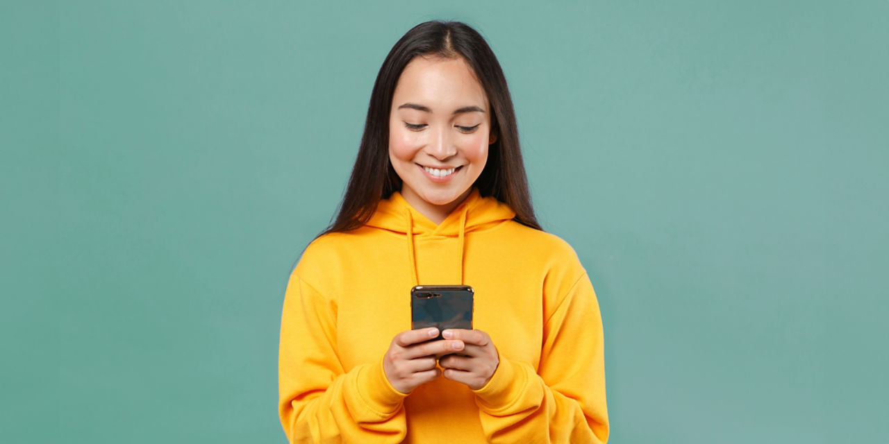Junge Frau mit schwarzen langen Haaren und gelbem Kapuzenpulli sieht lächelnd in ihr Smartphone, das sie mit beiden Händen hält.