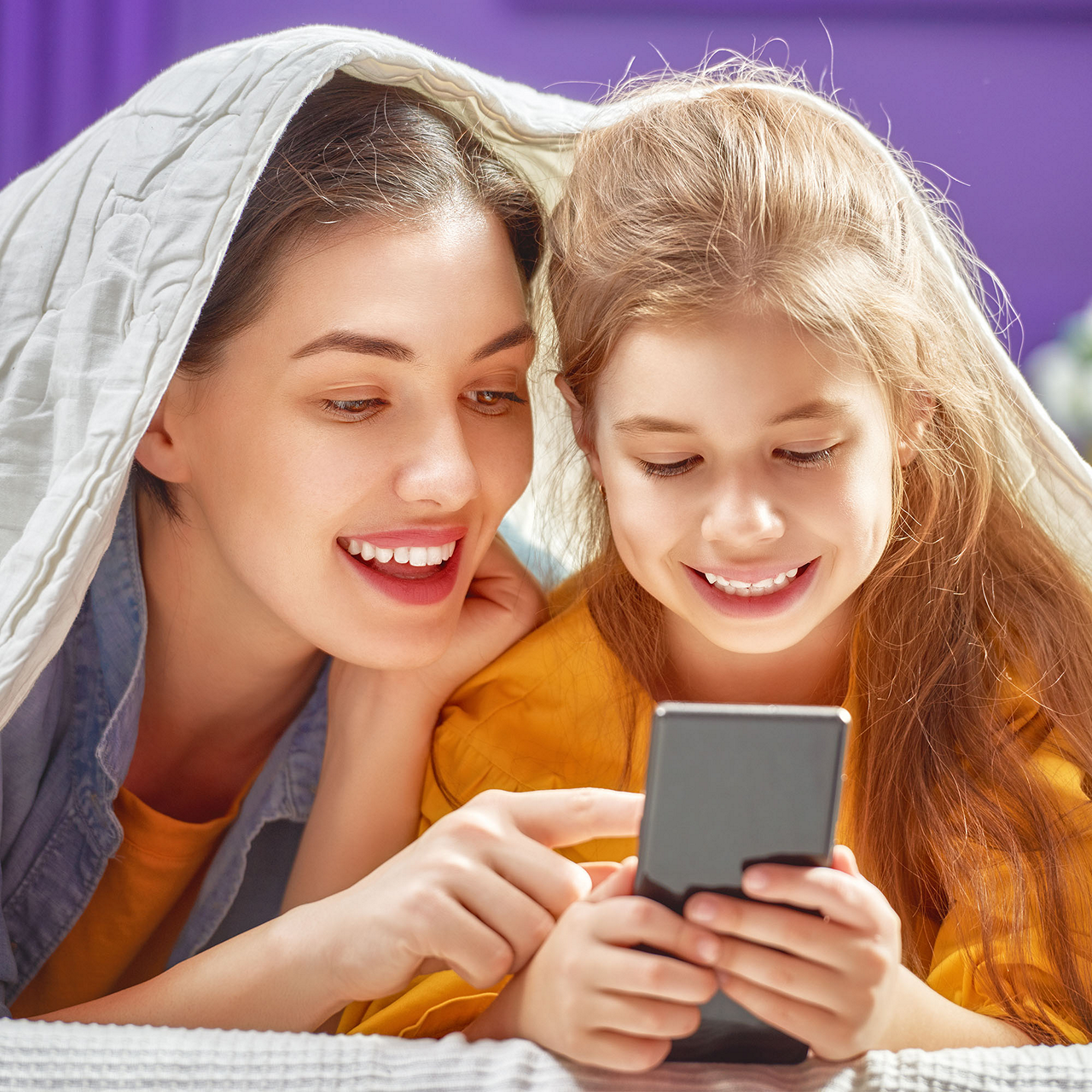 Mutter und Tochter sind unter einer Decke und lachen zusammen am Smartphone