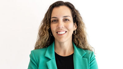 L'image montre un portrait professionnel d'une femme aux cheveux bruns mi-longs, souriante. Elle porte des boucles d’oreilles en perles et un haut noir, devant un fond doux et flou qui met en valeur son visage. L’environnement semble lumineux, indiquant un bon éclairage.