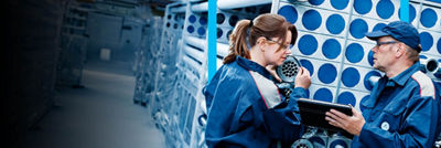 Two factory workers in blue uniforms inspecting equipment, one holding a tablet, in a highly industrial setting.