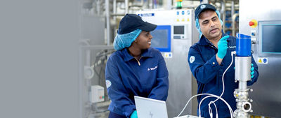 Dois trabalhadores da fábrica com uniformes azuis e redes de cabelo colaborando em um equipamento. Um deles explica seu funcionamento enquanto o outro ouve e trabalha em um laptop em um ambiente industrial de última geração.