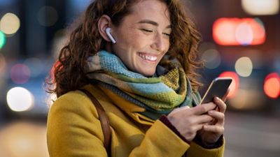 A woman with curly hair is wearing a yellow coat and a colourful scarf. She has earbuds in and is smiling while looking at her smartphone. The background is blurred, indicating an evening or night setting with city lights.