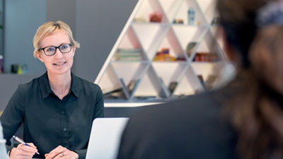 A woman with short blonde hair and glasses, wearing a black shirt, is engaged in a conversation. She is holding a pen and has a notebook and laptop in front of her. The background shows a modern office setting with geometric shelving.